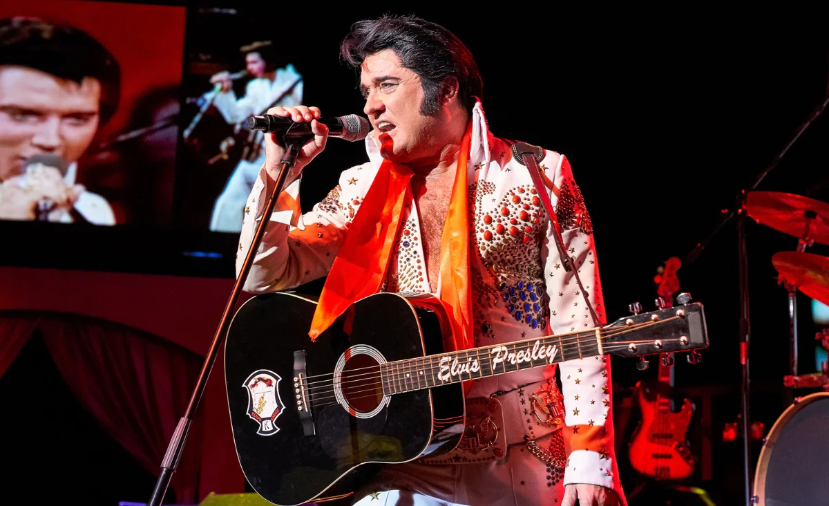Ein Künstler steht auf einer Bühne und hält ein Mikrofon in der rechten Hand. Er trägt ein auffälliges, mit Strasssteinen besetztes Bühnenkostüm im Stil von Elvis Presley sowie einen orangefarbenen Schal. Eine schwarze Gitarre mit dem Schriftzug 'Elvis Presley' und einem Wappenmotiv hängt vor seinem Körper. Im Hintergrund sind weitere Musikinstrumente und eine große Videoleinwand mit einer Bühnenaufnahme zu sehen. Die Szene ist farbenfroh ausgeleuchtet und vermittelt den Eindruck einer Live-Show. Der Kontext weist auf die Veranstaltung 'Stars in Concert' im Estrel Berlin hin.