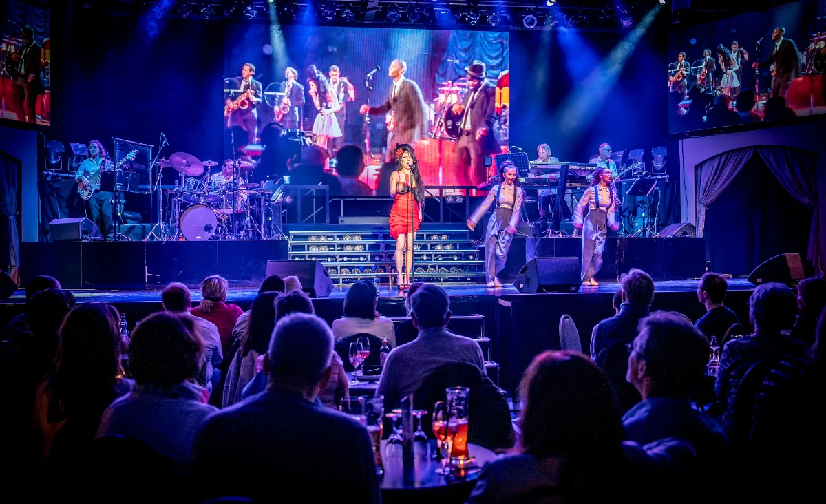 Bühne mit mehreren Musikerinnen und Musikern sowie einer Sängerin im Vordergrund, die in einem roten Kleid steht. Zwei Tänzerinnen mit auffälligen Kopfbedeckungen flankieren sie. Im Hintergrund sind weitere Bandmitglieder an Instrumenten zu sehen. Über der Bühne hängt eine große Videoleinwand, die die Bühnenszene in Echtzeit zeigt. Im Vordergrund sitzen zahlreiche Zuschauerinnen und Zuschauer an runden Tischen mit Getränken. Die Beleuchtung ist bunt und betont die Showatmosphäre. Die Szene spielt im Estrel in Berlin während der Live-Show »Stars in Concert«.