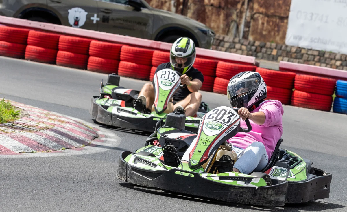 Zwei Personen in Helmen fahren in grünen Go-Karts auf einer Rennstrecke mit rot-blau-weißen Reifenbarrieren
