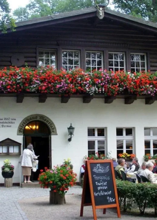 Im Vordergrund steht das historische Wirtshaus Moorlake mit weißer Fassade und dunklem Holzgiebel. Vor dem Gebäude befinden sich mehrere Tische und Stühle, an denen Gäste sitzen. Eine große, helle Sonnenschirm steht zentral auf der Terrasse. Links neben dem Eingang hängt ein Schild mit dem Namen des Wirtshauses, rechts davon steht eine schwarze Tafel mit weißer Schrift. Die Szene ist von üppigem, grünem Laub eingerahmt