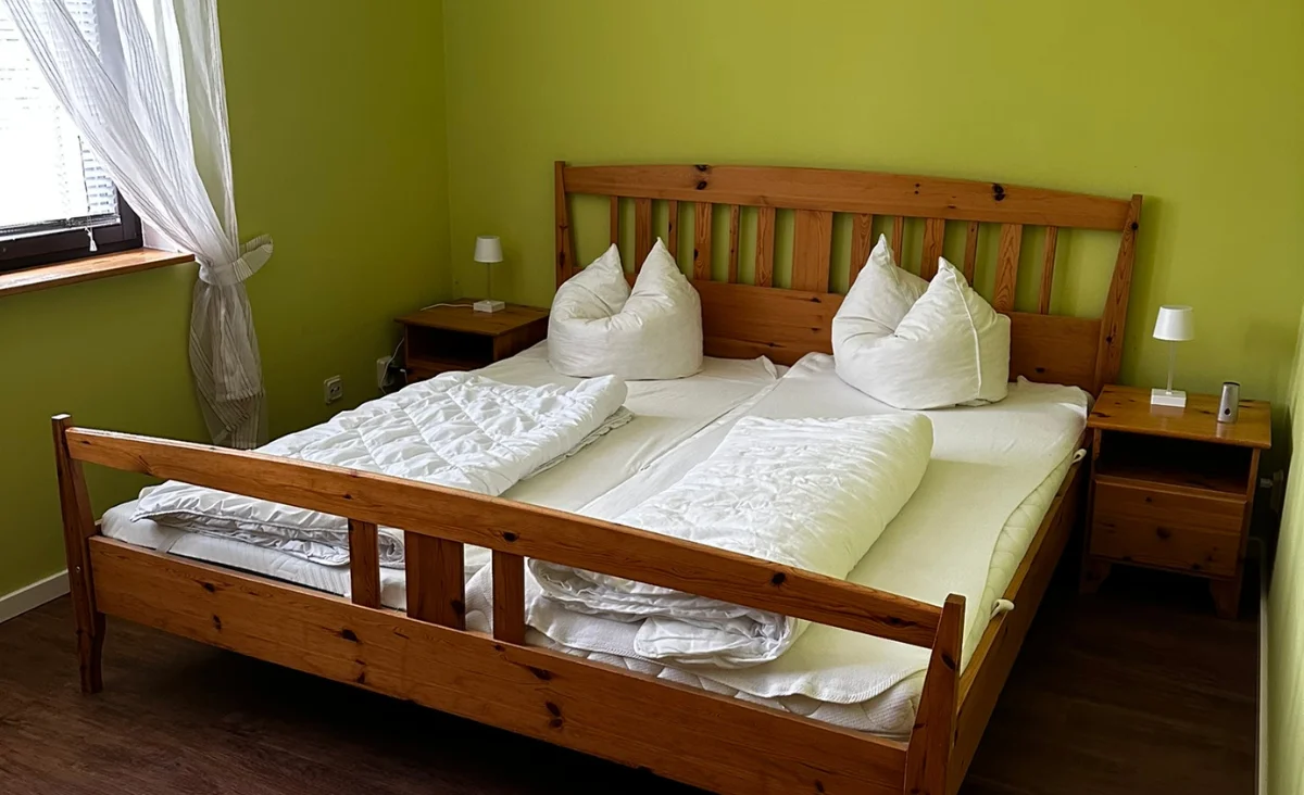 Holz-Doppelbett mit zwei weißen Kissen und Decken in einem Zimmer mit grün gestrichener Wand und zwei Nachttischen mit kleinen Lampen