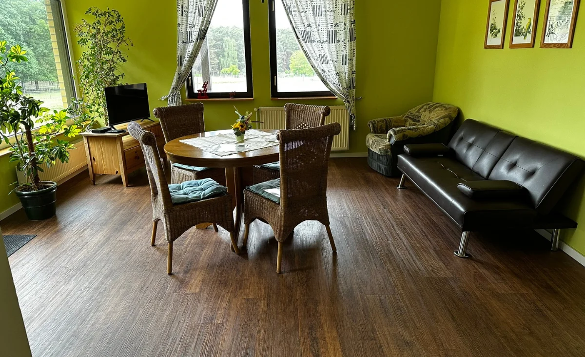 Wohnzimmer der Ferienwohnung im Wildpark Johannismühle mit grün gestrichenen Wänden, rundem Holztisch mit vier geflochtenen Stühlen, schwarzem Ledersofa, gemustertem Sessel, zwei Fenstern mit gemusterten Gardinen und einem kleinen Fernseher auf einem Holzschrank