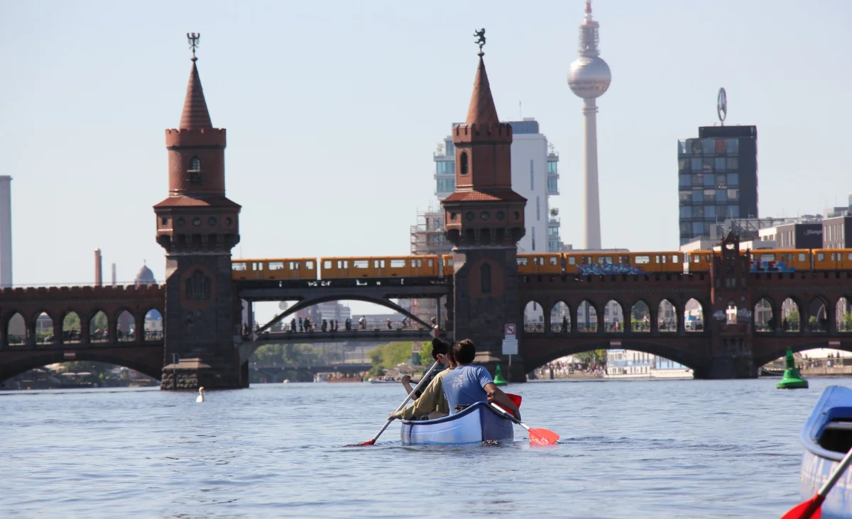 Zwei Personen paddeln in einem Kanu auf einem Fluss mit Blick auf die Oberbaumbrücke und die Berliner Fernsehturm im Hintergrund. Das Bild illustriert Kanutrail Berlin Landwehrkanal.