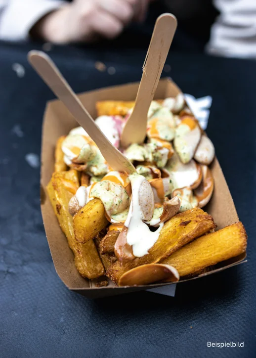 In einer braunen Pappschale liegen dicke, goldgelbe Pommes frites, die mit verschiedenen Saucen und kleinen Würstchenscheiben belegt sind. Zwei Holzspatel stecken in der Portion. Im Hintergrund ist eine Person unscharf zu erkennen. Die Aufnahme entstand auf dem Street Food Festival Ludwigsfelde.