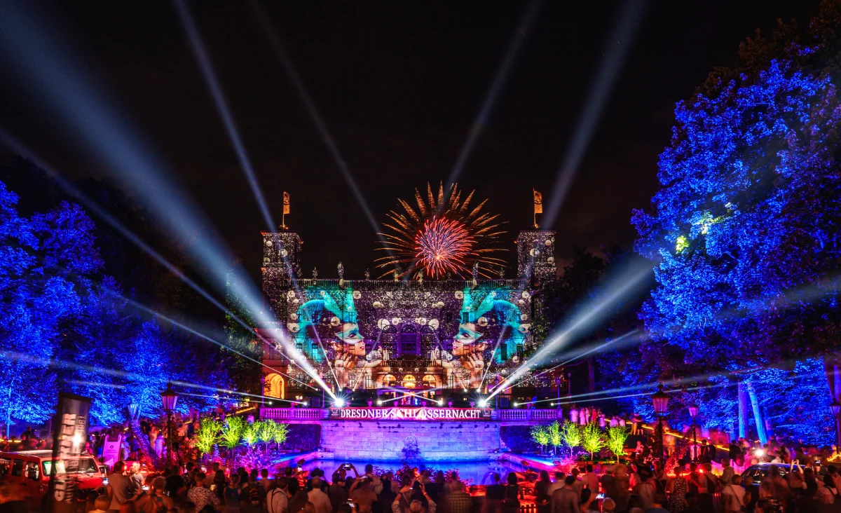Feuerwerk und Lichtstrahlen über einem historischen Schloss mit beleuchteten Bäumen und einer Menschenmenge bei der Dresdner Schlössernacht.