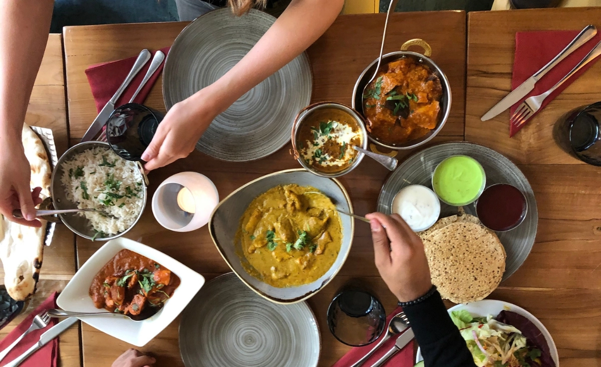 Tisch mit indischen Gerichten, darunter Reis, Curry, Saucen und Papadam, Personen greifen mit Löffeln und Händen zu.