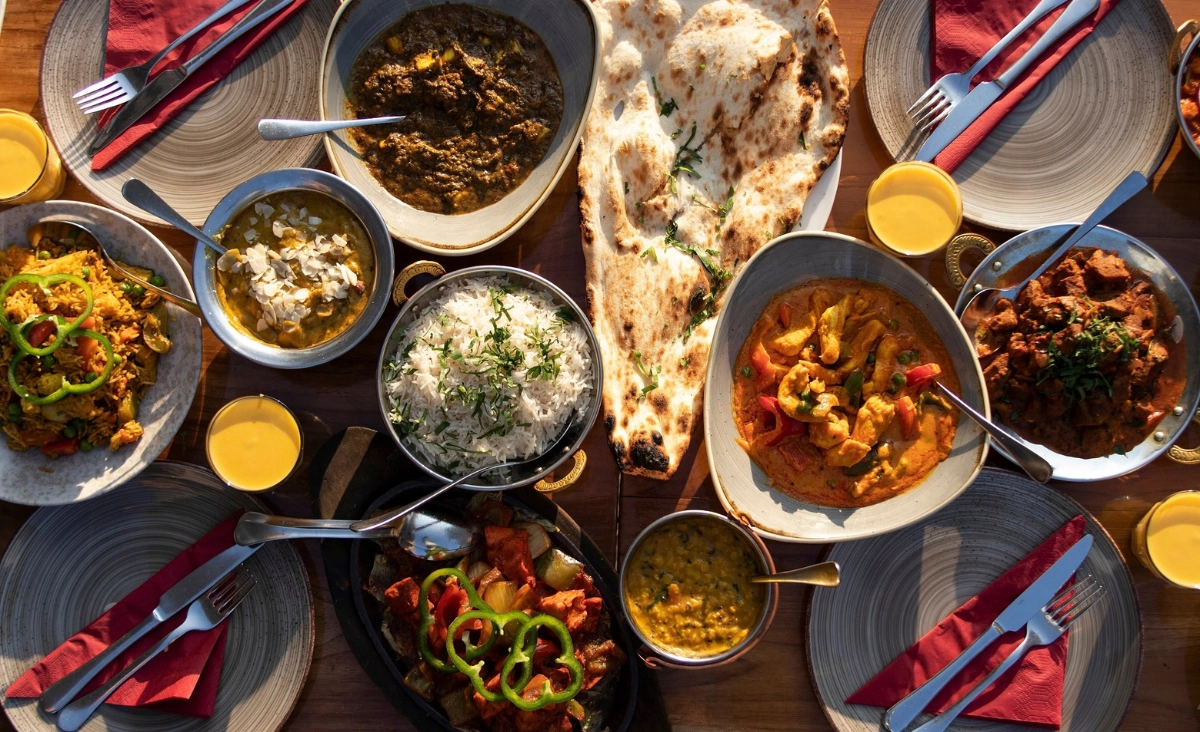 Tisch mit indischen Gerichten, darunter Naan-Brot, Reis, verschiedene Currys und Linsengerichte, umgeben von Tellern mit Besteck und Gläsern mit Orangensaft.