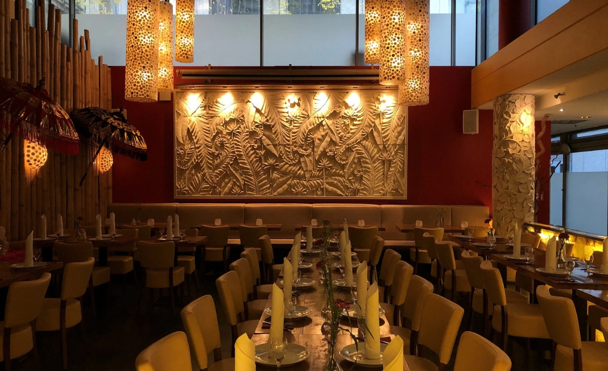 Innenraum eines Restaurants mit langen Tischen, gedeckten Plätzen, dekorativer Wand mit Pflanzenrelief und warmen Hängeleuchten.