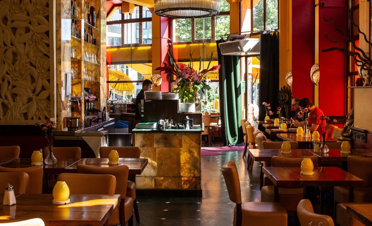 Innenraum eines Restaurants mit Holztischen, bequemen Stühlen, roten Wänden, großen Fenstern und einer Theke in der Mitte.
