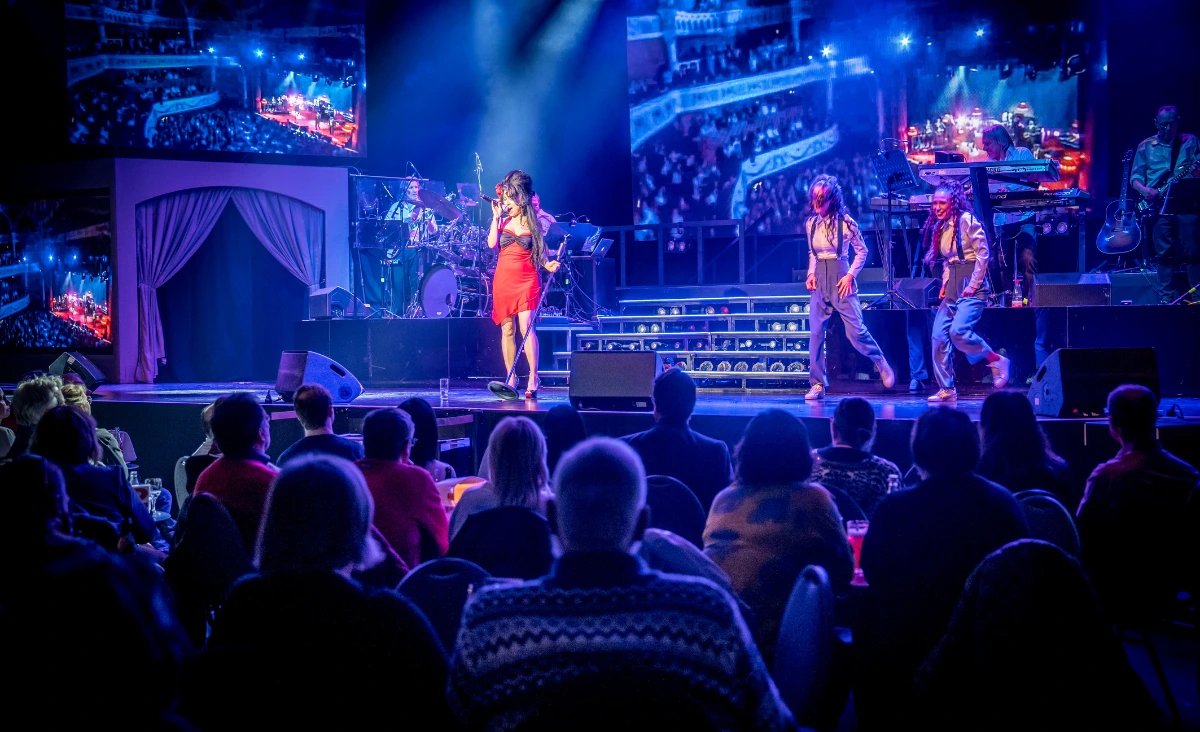 Sängerin in rotem Kleid und schwarzer Perücke mit Hochsteckfrisur steht auf Bühne vor Band und Publikum im Estrel Showtheater Berlin bei Amy Winehouse Tribute Show