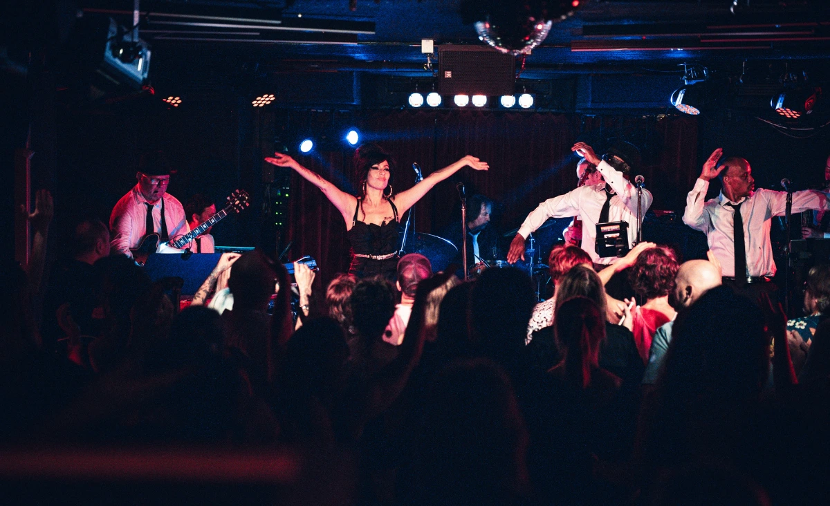 Sängerin in schwarzem Kleid und schwarzer Perücke mit Hochsteckfrisur steht auf Bühne vor Band und Publikum im Estrel Showtheater Berlin bei Amy Winehouse Tribute Show