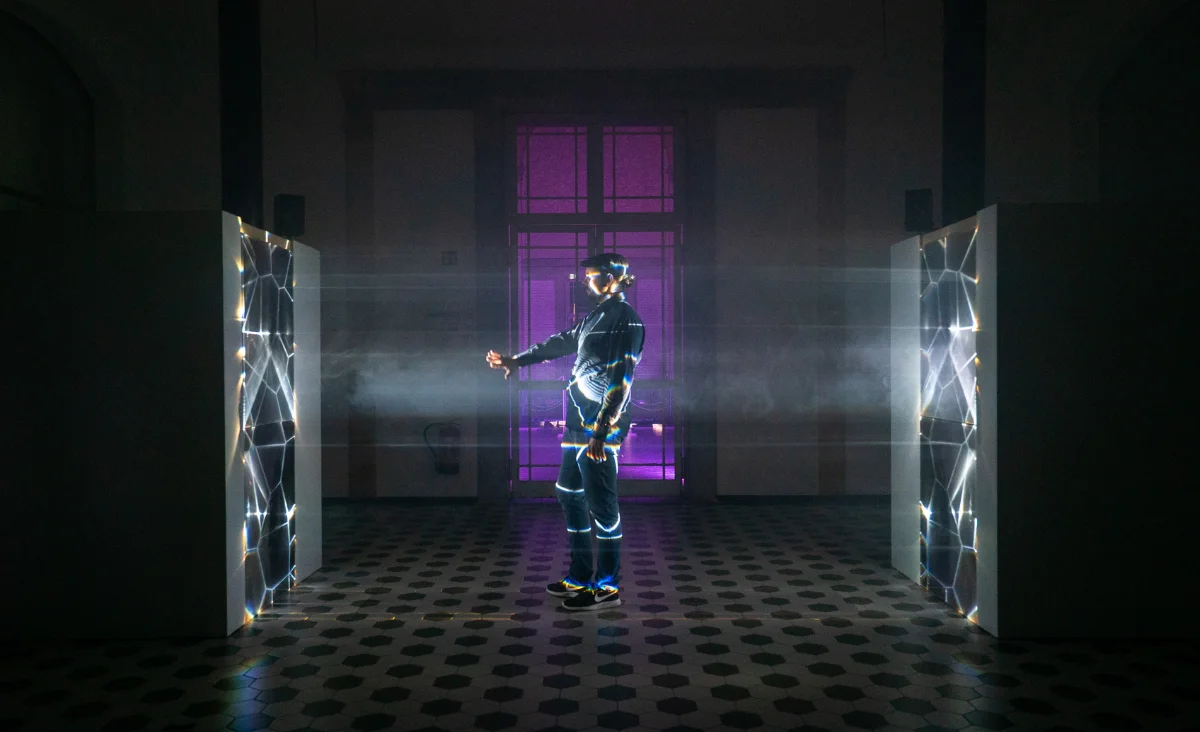 Person mit Virtual-Reality-Brille steht zwischen zwei beleuchteten, netzartigen Installationen in einem dunklen Raum mit Fenster im Hintergrund