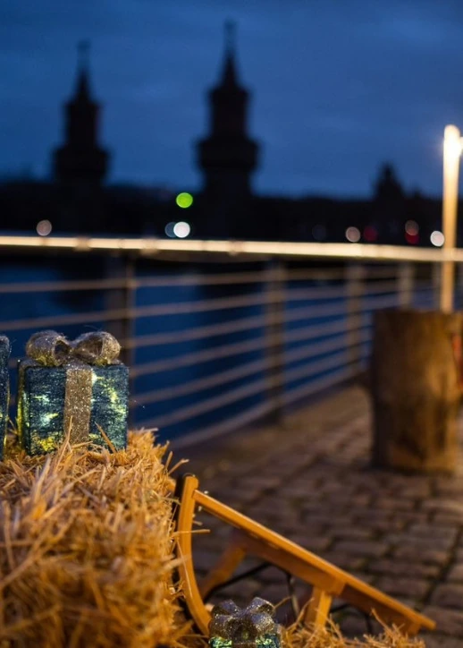Detailansicht einer winterlichen Dekoration auf dem All inclusive Weihnachtsmarkt an der Spree in Berlin: Zwei glitzernde Geschenke auf einem Strohballen im Vordergrund, mit der beleuchteten Oberbaumbrücke und dem Spreespeicher im Hintergrund am Abend.