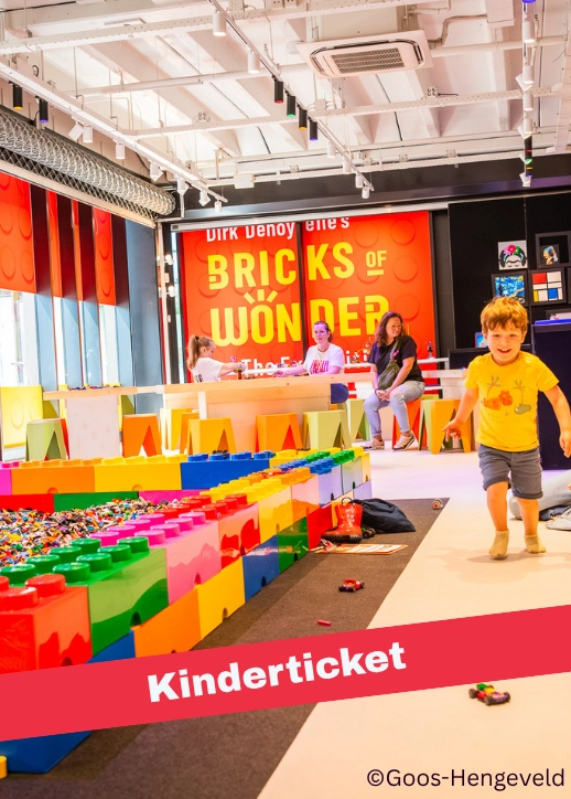 Ein Kind läuft auf einem hellen Boden vor einer Frau sitzend neben einem großen, bunten Lego-Bau mit großen Steinen in einem Ausstellungsraum von Bricks of Wonders.