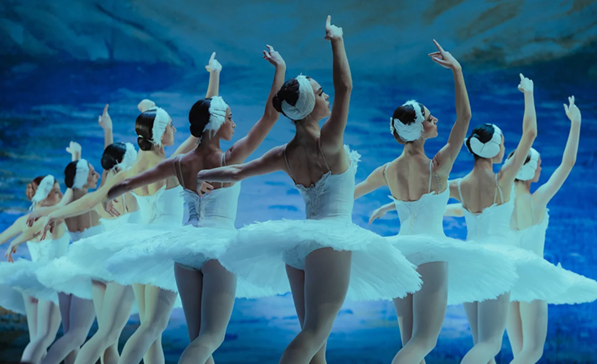 Gruppe von Ballerinas in weißen Tütü und Kopfschmuck, die in einer Linie mit erhobenen Armen tanzen vor blauem Himmel-Hintergrund