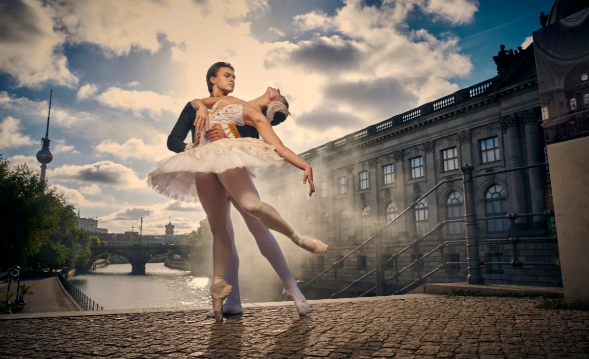 Zwei Balletttänzer in klassischer Pose vor einem historischen Gebäude und einem Fluss unter bewölktem Himmel