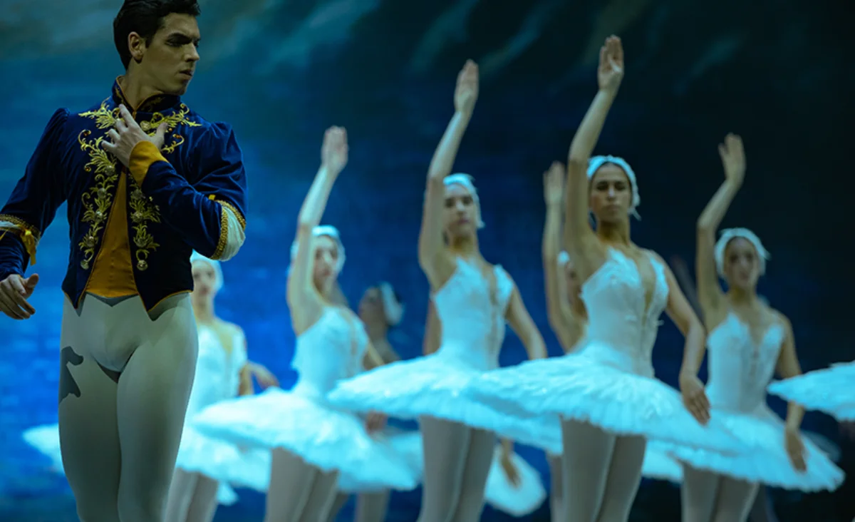 Ein männlicher Balletttänzer in blau-goldener Jacke und weißer Hose steht links, im Hintergrund eine Gruppe weiblicher Tänzerinnen in weißen Tütü mit erhobenen Armen.